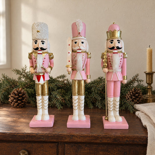 I tre schiaccianoci rosa confetto e oro in fila su una credenza di legno, fra rami di abete, pigne e un candeliere